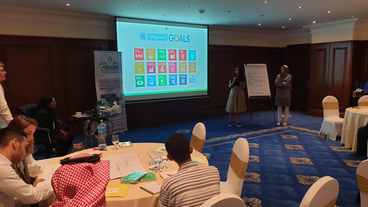 Piozone facilitators leading a workshop on the UN Sustainable Development Goals, with the seventeen goal tiles projected on screen and participants working in groups at round tables